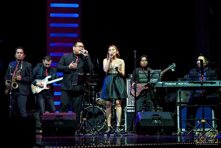 J-Project performing live on a concert stage with dual lead vocalists, saxophone, electric guitar, bass, keyboards and drums